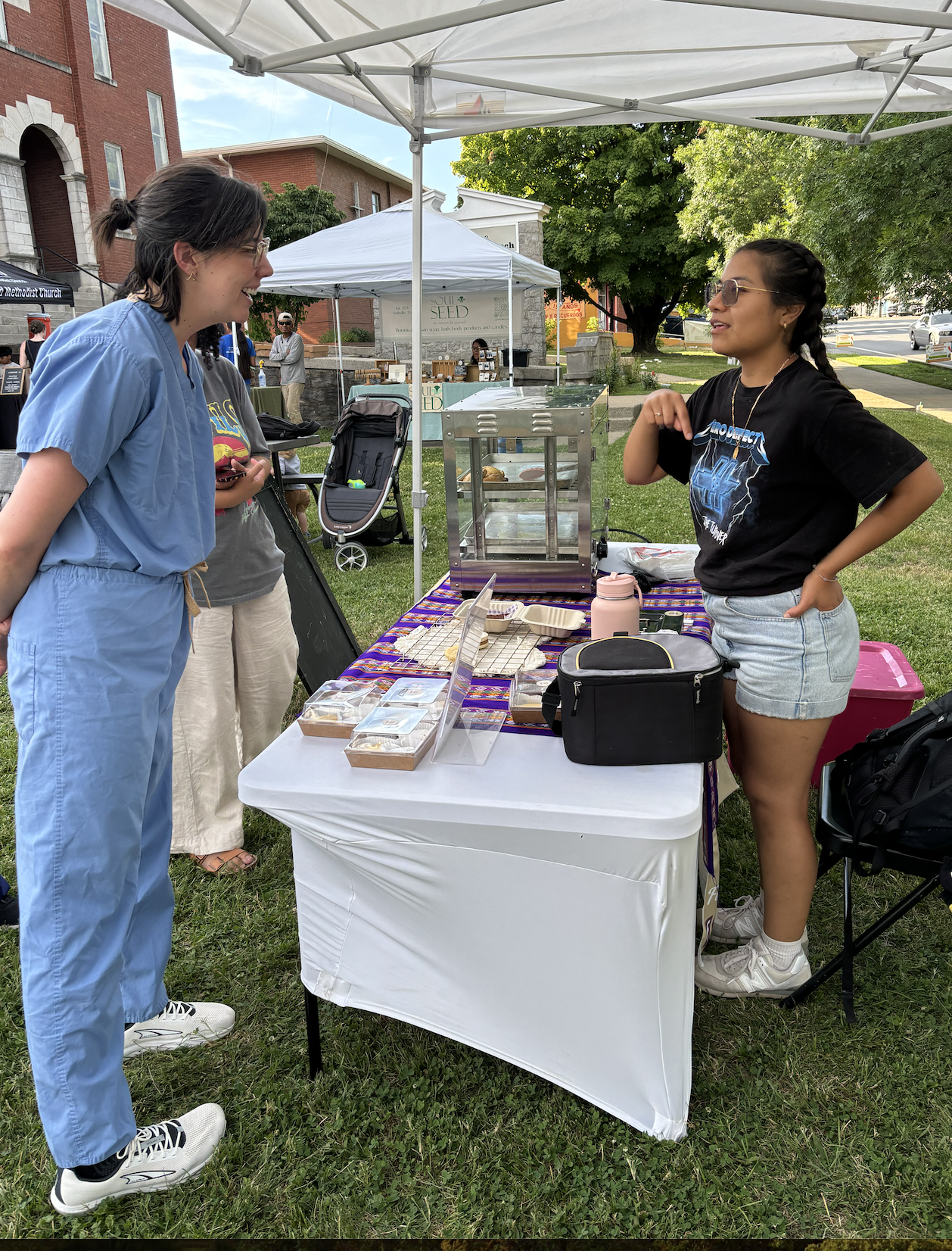 A woman dressed in scrubs at a farmer's market talks to a Latina woman selling empanadas